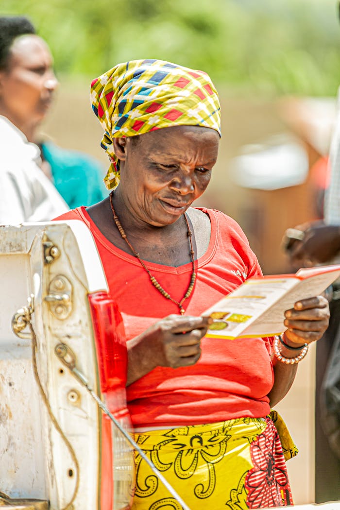 Elderly woman wearing headscarf reads a leaflet outdoors in sunny setting.